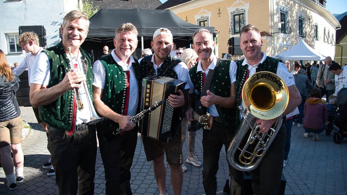 Eine Musikgruppe mit fünf Männern in traditionellen Trachten steht lächelnd zusammen. Sie halten verschiedene Instrumente und befinden sich auf einer Veranstaltung im Freien. | © Echtzeit TV