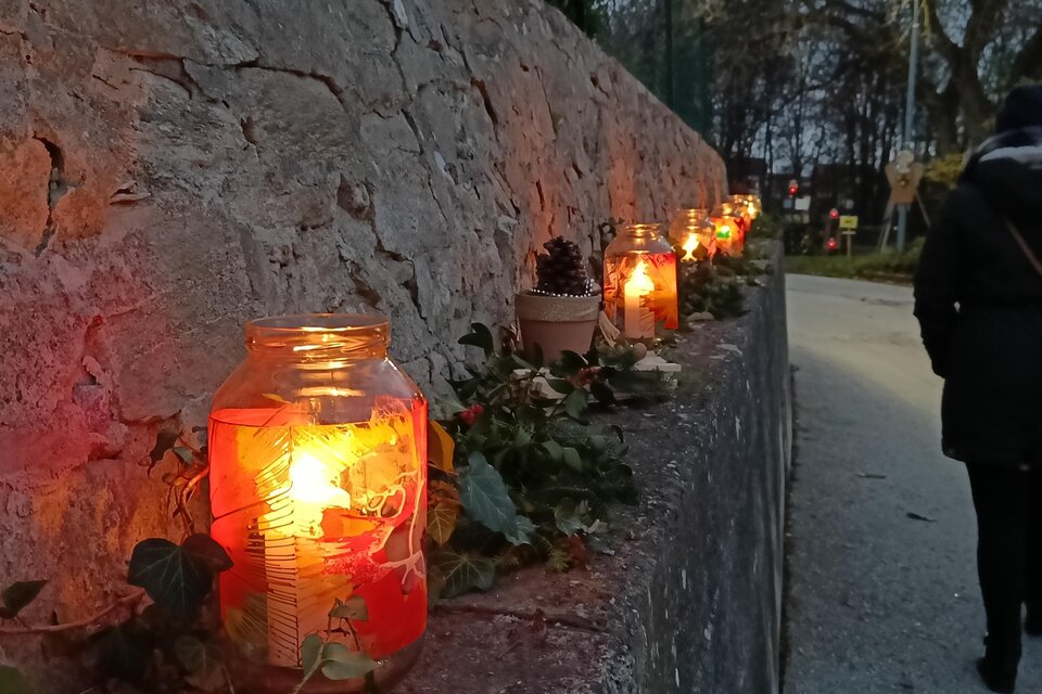 Ein Weg mit einer Steinmauer, dekoriert mit leuchtenden Gläsern, die Kerzen enthalten. Im Hintergrund ist eine Person zu sehen, die vorbeigeht, während die Dämmerung einsetzt. | © Kreativteam Adventpfad Pöllau