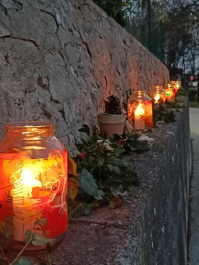 Egy út egy kőfal mentén, fényes üvegekkel díszítve, amelyek gyertyákat tartalmaznak. A háttérben egy személy látható, aki elhalad, miközben beáll a szürkület. | © Kreativteam Adventpfad Pöllau