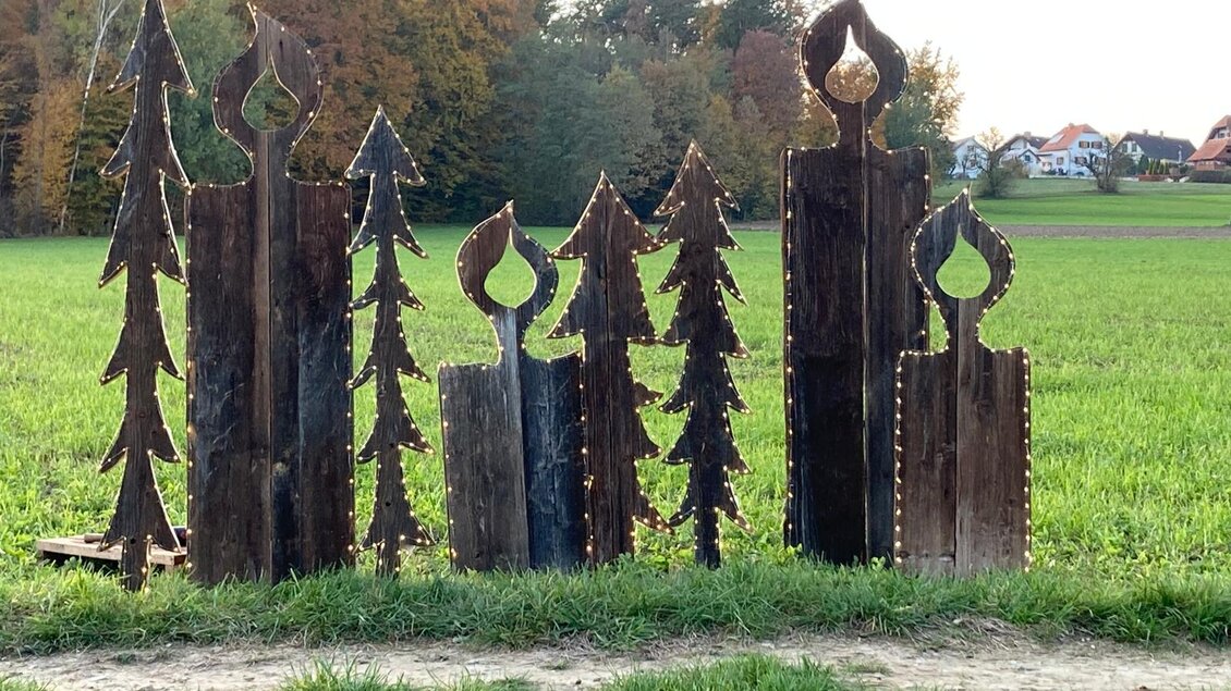 Eine Gruppe von Holzskulpturen in Form von Bäumen und Kerzen steht auf einem Feld. Im Hintergrund sind sanfte Hügel und Wohnhäuser sichtbar. | © Angela Geier