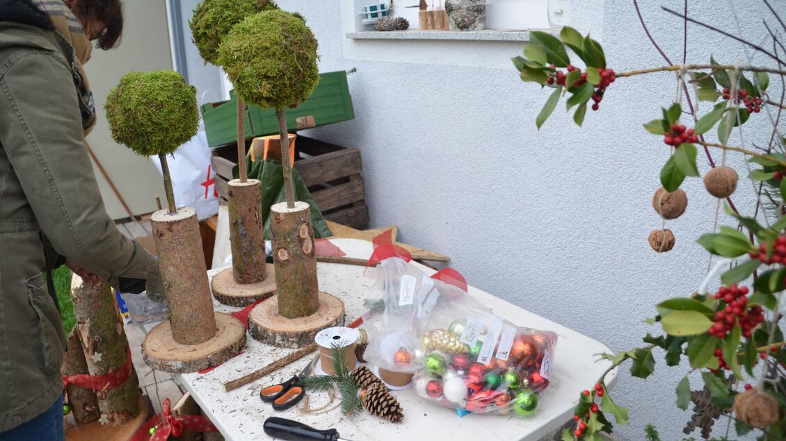 Eine Person gestaltet dekorative Pflanzen aus Moos und Holz auf einem Tisch. Es sind verschiedene Bastelmaterialien sowie Weihnachtsdekorationen sichtbar. | © Kreativteam Adventpfad Pöllau