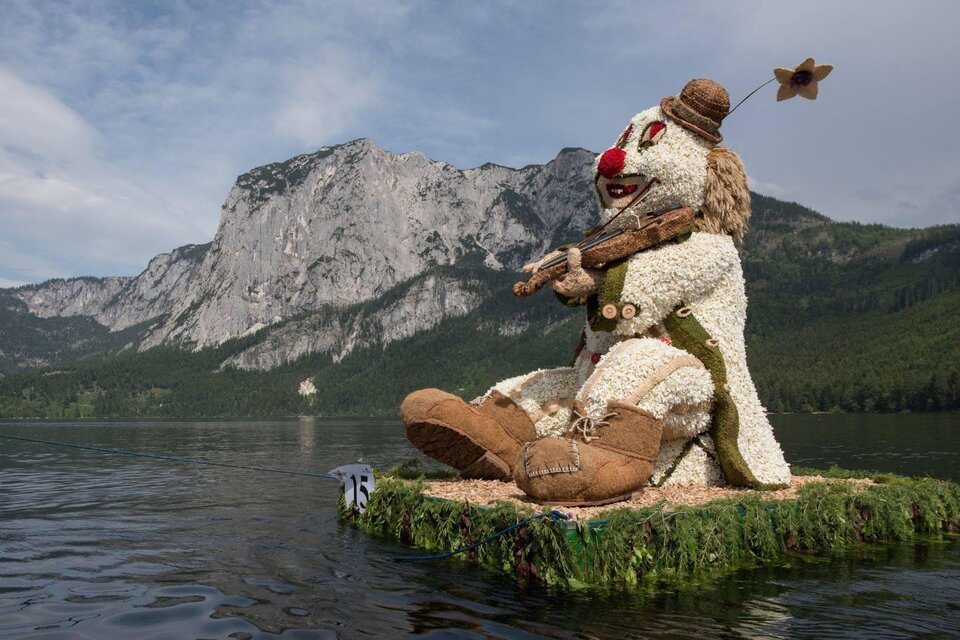 Eine große, aus Blumen und Naturmaterialien gestaltete Figur eines Clowns sitzt auf einem Boot im Wasser. Im Hintergrund sind majestätische Berge und ein klarer Himmel zu sehen. | © M.Baumgartner