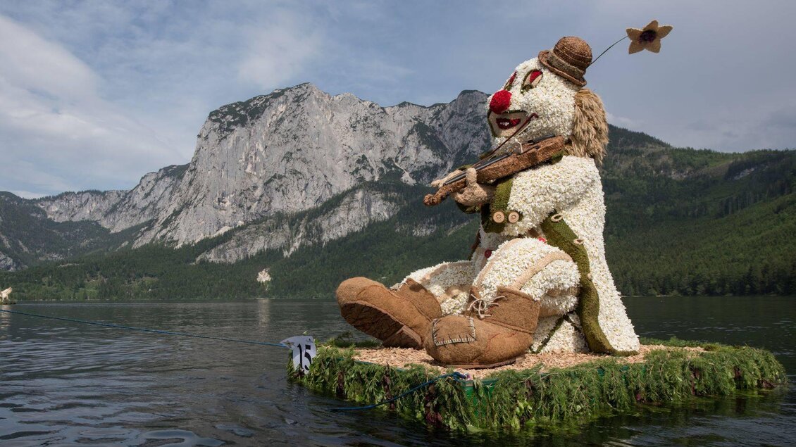 Eine große, aus Blumen und Naturmaterialien gestaltete Figur eines Clowns sitzt auf einem Boot im Wasser. Im Hintergrund sind majestätische Berge und ein klarer Himmel zu sehen. | © M.Baumgartner