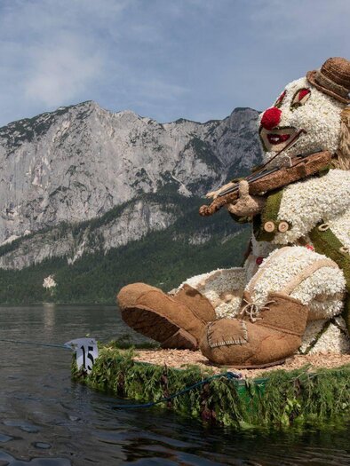 A large figure of a clown made of flowers and natural materials sits on a boat in the water. In the background, majestic mountains and a clear sky can be seen. | © M.Baumgartner