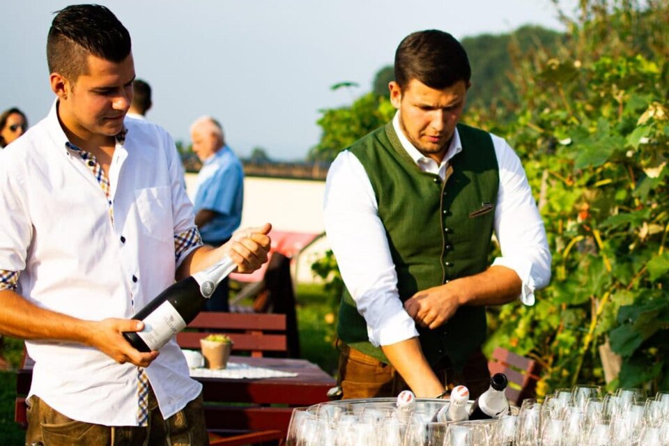 Zwei Männer stehen an einem Tisch mit Gläsern, während sie Schaumwein einschenken. Die Umgebung ist grün und sonnig, perfekt für eine Feier im Freien. | © Ratscher Landhaus GmbH