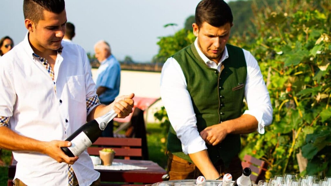 Zwei Männer stehen an einem Tisch mit Gläsern, während sie Schaumwein einschenken. Die Umgebung ist grün und sonnig, perfekt für eine Feier im Freien. | © Ratscher Landhaus GmbH
