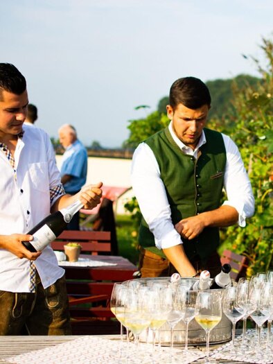 Two men are standing at a table with glasses while pouring sparkling wine. The surroundings are green and sunny, perfect for an outdoor celebration. | © Ratscher Landhaus GmbH