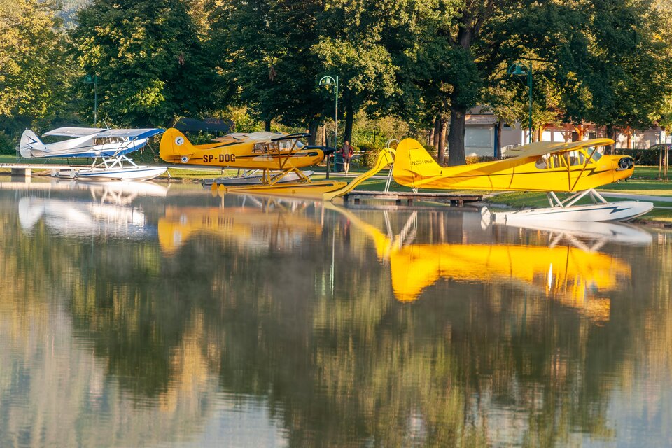 Bunte Wasserflugzeuge liegen am Ufer eines ruhigen Sees. Die Reflexionen der Flugzeuge sind auf der Wasseroberfläche zu sehen. | © Chris Barszczewski