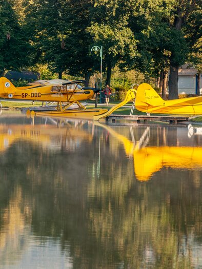 Colorful seaplanes rest on the shore of a calm lake. The reflections of the planes can be seen on the water's surface. | © Chris Barszczewski