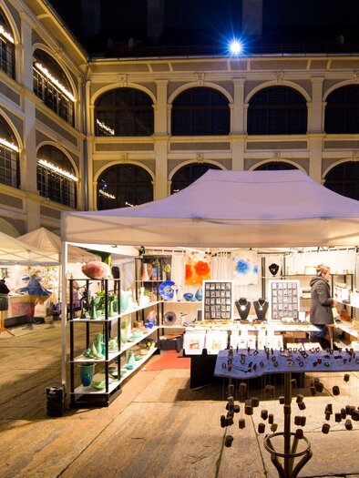 A night market with white tents and colorful stalls. Visitors view handcrafted artworks and decorations. | © graz toursimus- -Harry Schiffer