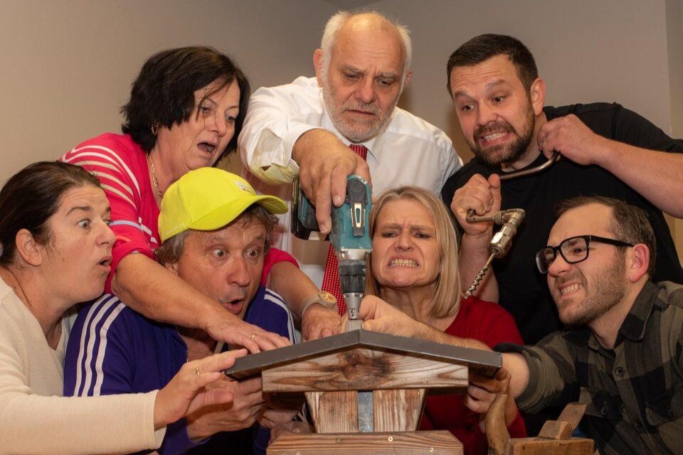 Eine Gruppe von Menschen schaut gespannt auf ein Handwerksprojekt. Sie halten Werkzeuge und zeigen verschiedene Emotionen, während sie zusammenarbeiten. | © Komödienbühne Gosdorf