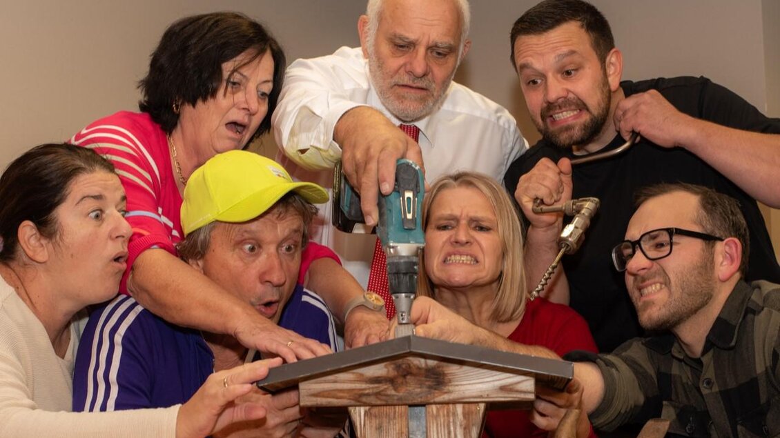 Eine Gruppe von Menschen schaut gespannt auf ein Handwerksprojekt. Sie halten Werkzeuge und zeigen verschiedene Emotionen, während sie zusammenarbeiten. | © Komödienbühne Gosdorf