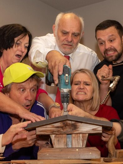 Eine Gruppe von Menschen schaut gespannt auf ein Handwerksprojekt. Sie halten Werkzeuge und zeigen verschiedene Emotionen, während sie zusammenarbeiten. | © Komödienbühne Gosdorf
