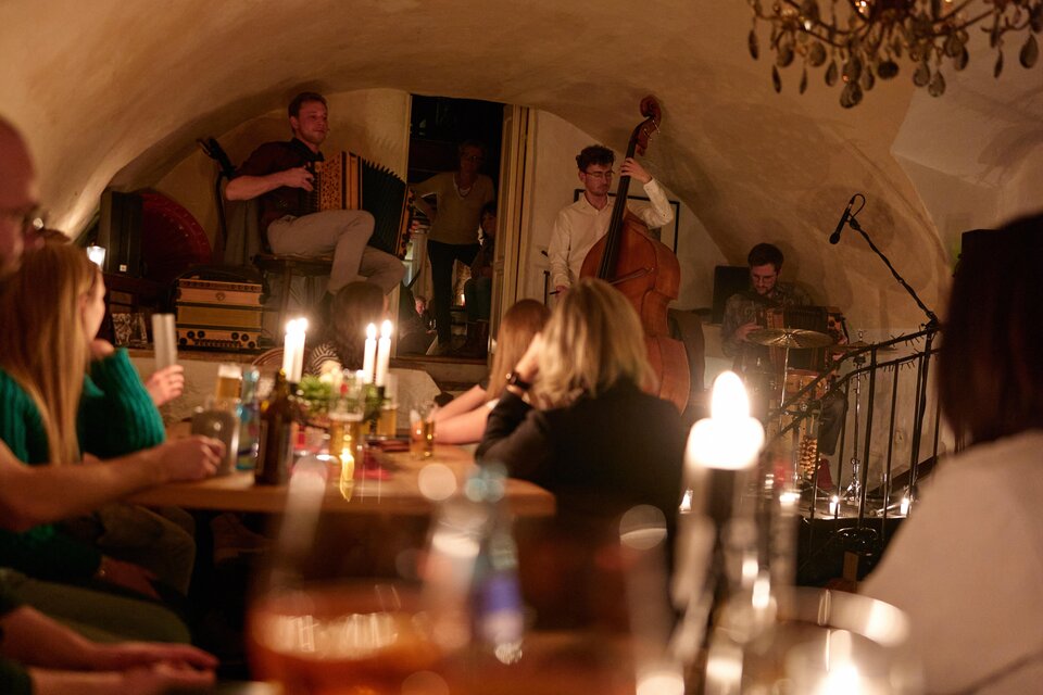 Eine gemütliche Abendveranstaltung in einem Keller mit Live-Musik. Musiker spielen auf der Bühne, während das Publikum an Tischen sitzt und die Atmosphäre genießt. | © Bernd Wimmer