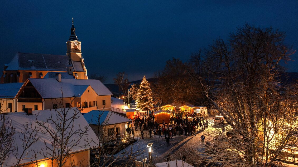 Eine winterliche Szene mit einem festlich beleuchteten Weihnachtsmarkt. Schnee bedeckt die Dächer, und Menschen versammeln sich um die Stände. | © Gemeinde Pöllauberg