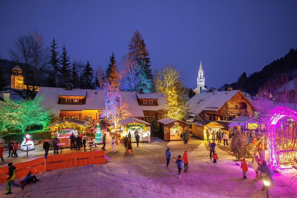 Ein festlicher Weihnachtsmarkt bei Nacht, erleuchtet von bunten Lichtern. Menschen genießen die festliche Stimmung um ein geschmücktes Dorf. | © Martin Huber