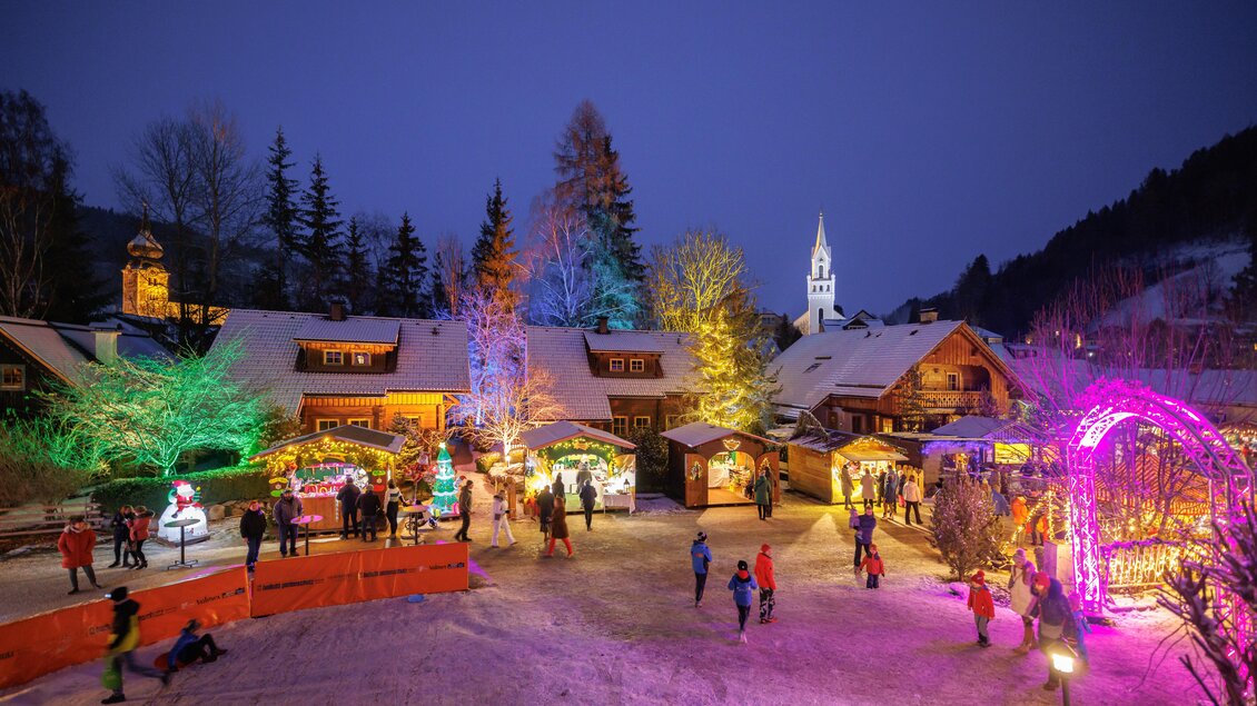 Ein festlicher Weihnachtsmarkt bei Nacht, erleuchtet von bunten Lichtern. Menschen genießen die festliche Stimmung um ein geschmücktes Dorf. | © Martin Huber
