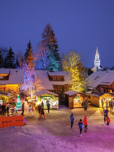 Ein festlicher Weihnachtsmarkt bei Nacht, erleuchtet von bunten Lichtern. Menschen genießen die festliche Stimmung um ein geschmücktes Dorf. | © Martin Huber