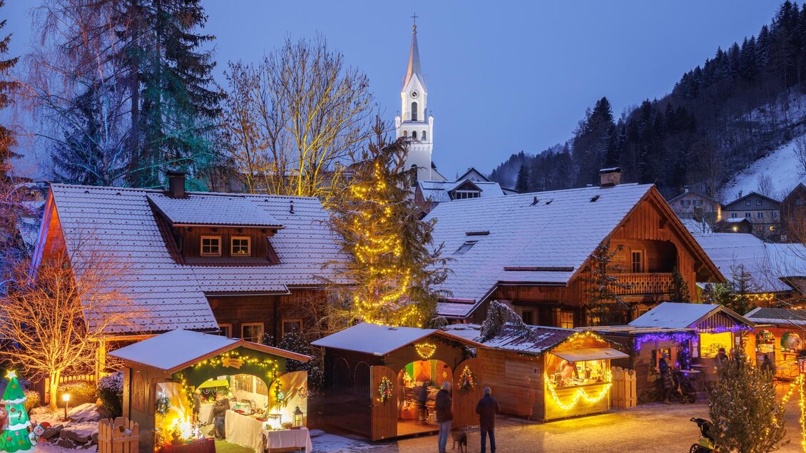 Ein malerisches Winterdorf mit verschneiten Dächern und festlich geschmückten Buden. Im Hintergrund steht eine Kirche mit einem hohen Turm. | © Martin Huber