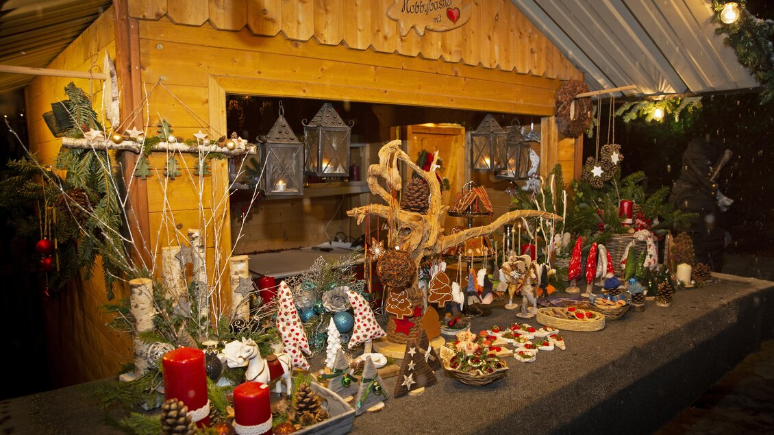 Ein festlich dekorierter Weihnachtsstand aus Holz mit einer Vielzahl von handgemachten Leckereien und Dekorationen. Kerzen, Tannenzweige und geflochtene Elemente schaffen eine winterliche Atmosphäre. | © Herbert Raffalt