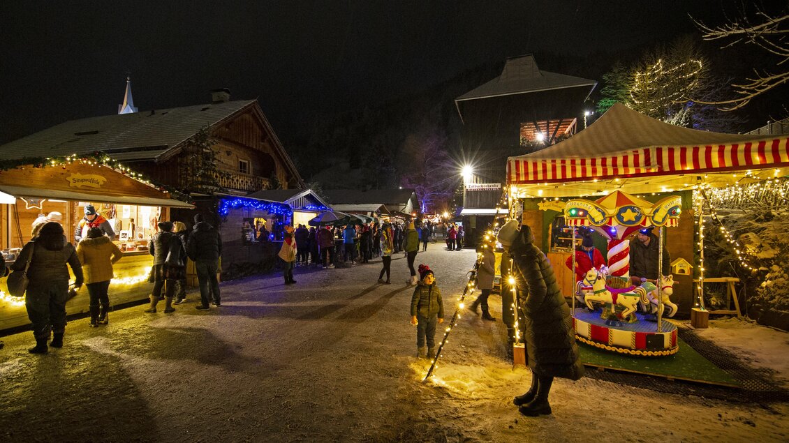 Ein festlicher Weihnachtsmarkt bei Nacht mit bunten Lichtern und Ständen. Menschen flanieren über den Schneebedeckten Platz und genießen die Atmosphäre. | © TVB Schladming-Dachstein