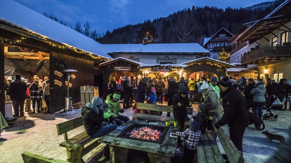 Ein Weihnachtsmarkt mit vielen Menschen, die um ein Feuer sitzen. Die festlich beleuchteten Stände und ein winterlicher Hintergrund schaffen eine gemütliche Atmosphäre. | © Gerhard Pilz