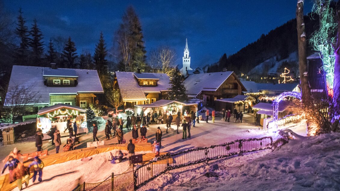 Eine festliche Winterlandschaft mit geschmückten Hütten und Besuchern auf einem verschneiten Platz. Im Hintergrund leuchtet ein Kirchturm in der Nacht. | © Gerhard Pilz