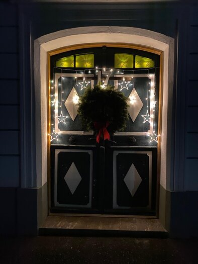 Eine festlich dekorierte Eingangstür mit Lichtern und einem Kranz. Die Tür ist in dunklem Holz gehalten und wird von einem warmen Lichtschein erleuchtet. | © Alexandra Wippel