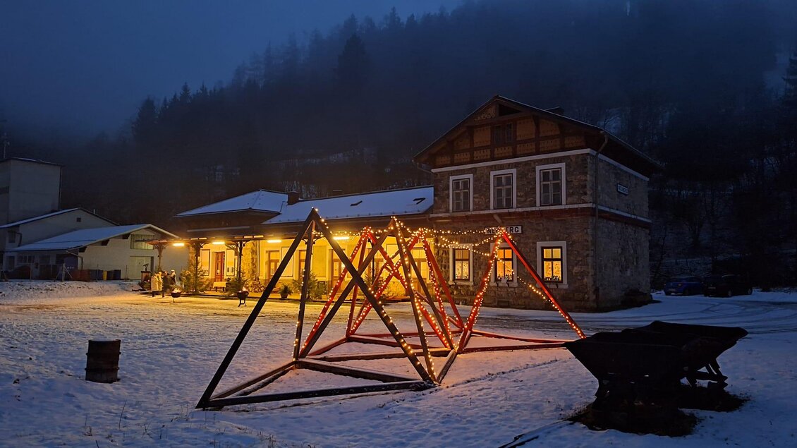 Ein winterliches Dorf bei Nacht mit einem beleuchteten Spielplatz und einem alten Gebäude im Hintergrund. Der Schnee reflektiert das Licht und schafft eine ruhige Atmosphäre. | © Bahnhof Neuberg, Paier