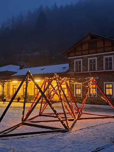 A winter village at night with a illuminated playground and an old building in the background. The snow reflects the light and creates a tranquil atmosphere. | © Bahnhof Neuberg, Paier