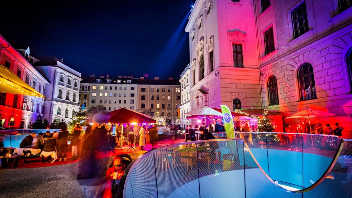 Eine lebhafte Nacht mit bunten Lichtern und einer festlichen Atmosphäre. Menschen genießen die Zeit in einem Außenbereich mit verschiedenen Ständen und beleuchteten Gebäuden. | © Graz Tourismus - Werner Krug