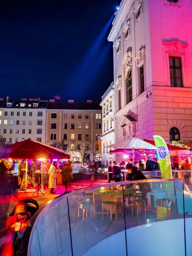 A lively night with colorful lights and a festive atmosphere.  
People enjoy their time in an outdoor area with various stalls and illuminated buildings. | © Graz Tourismus - Werner Krug