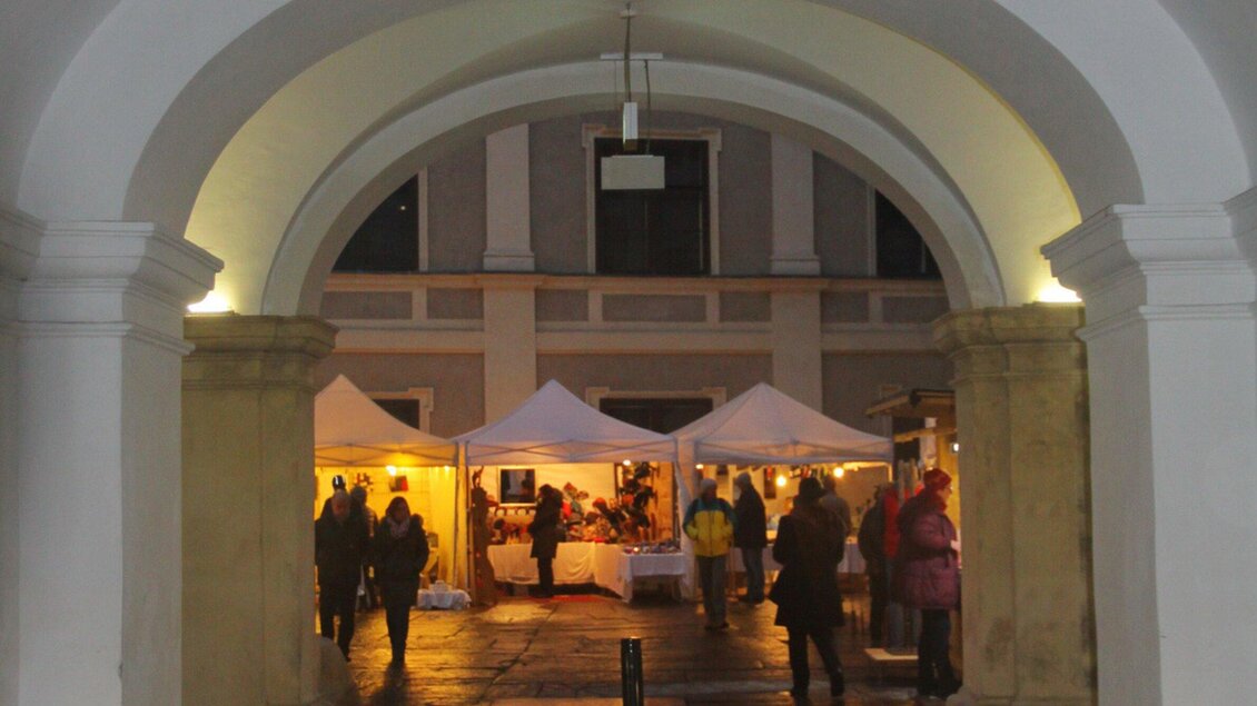 Ein Marktplatz mit festlich beleuchteten Ständen unter einem großen Bogen. Menschen schlendern durch die Gasse und genießen die Atmosphäre. | © Steirische Initiative Kunsthandwerk - Hans Walters