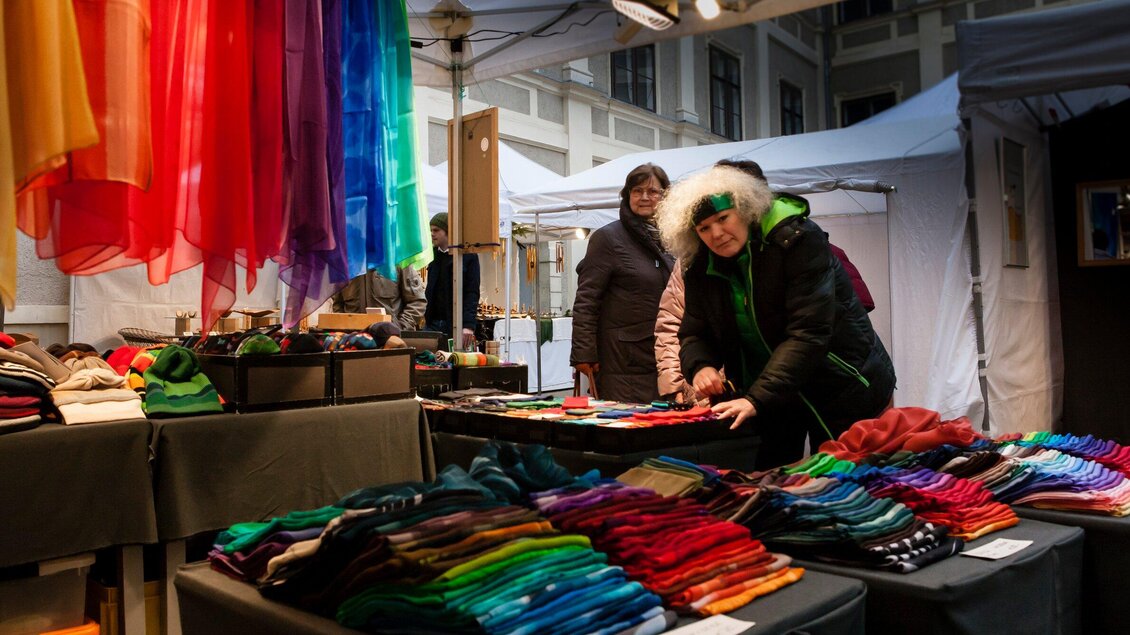Ein Marktstand mit bunten Tüchern und Schals. Zwei Frauen schauen sich die Waren an und lächeln. | © Steirische Initiative Kunsthandwerk - Nandor Vidak