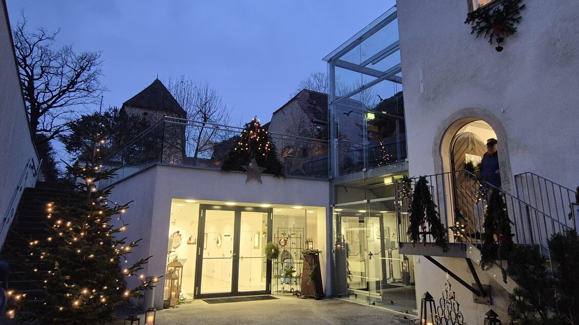 Ein einladender Eingang zu einem Gebäude mit Weihnachtsbeleuchtung und einem Christbaum. Der Himmel ist dämmerig und verleiht der Szenerie eine festliche Atmosphäre. | © Oststeiermark Tourismus