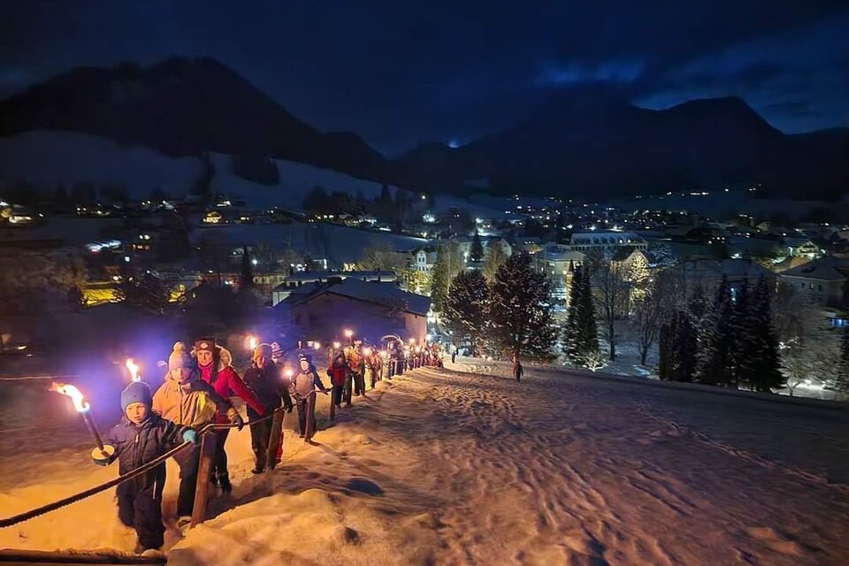 Eine Gruppe von Menschen trägt Fackeln und wandert durch eine schneebedeckte Landschaft. Im Hintergrund leuchtet eine kleine Stadt unter einem klaren Nachthimmel. | © Kirtagsverein