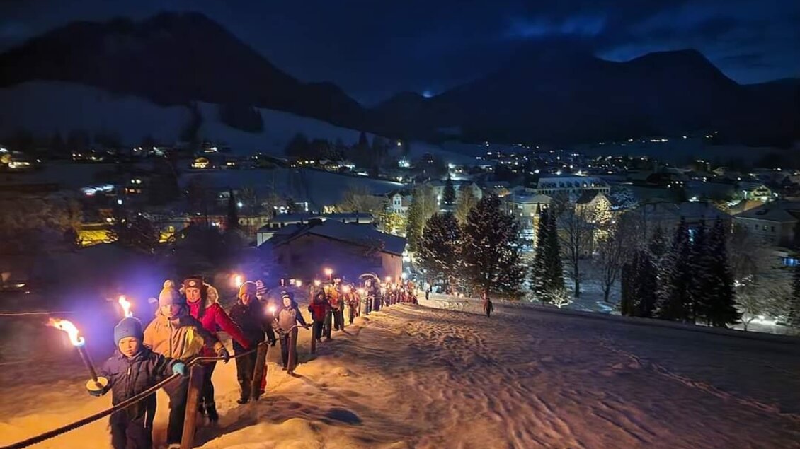 Eine Gruppe von Menschen trägt Fackeln und wandert durch eine schneebedeckte Landschaft. Im Hintergrund leuchtet eine kleine Stadt unter einem klaren Nachthimmel. | © Kirtagsverein