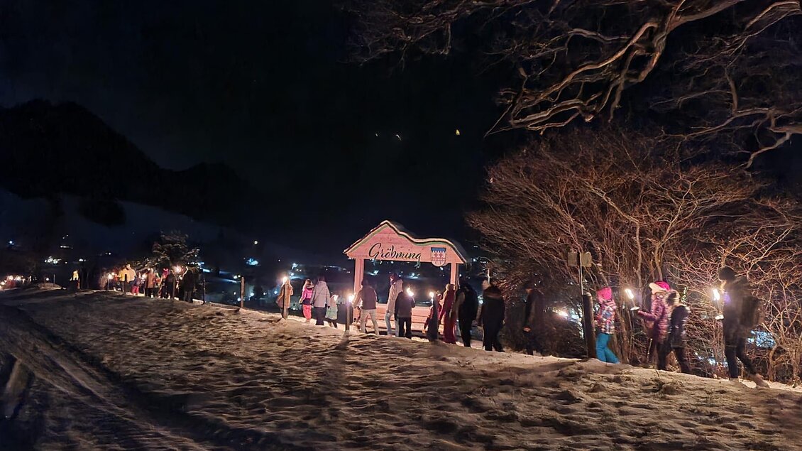 Eine Gruppe von Menschen wandert nachts mit Fackeln durch eine schneebedeckte Landschaft. Im Hintergrund sind Bäume und eine leuchtende Markierung sichtbar. | © Kirtagsverein