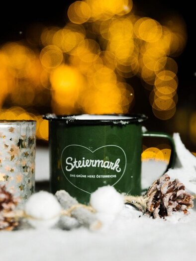 A green cup and a silver candle holder are on a snow-covered table. Surrounded by pine cones and a cozy light ambiance. | © Lupi Spuma