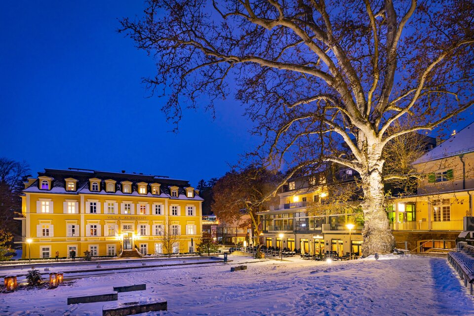 Eine malerische Winterlandschaft mit einem beleuchteten gelben Gebäude und einem großen Baum. Der Schnee bedeckt den Boden, und der Abendhimmel ist dunkelblau. | © Thermen- & Vulkanland