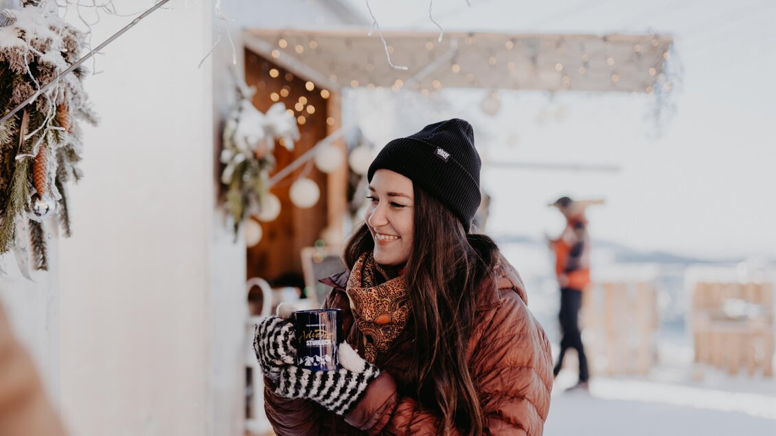 Eine junge Frau in warmen Winterkleidung lächelt und hält einen Becher in den Händen. Im Hintergrund sind festliche Lichter und Schnee zu sehen. | © nicoleseiser.at