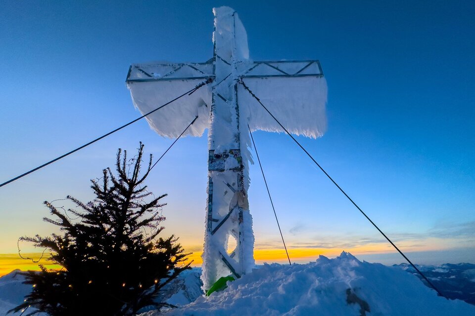 Ein großes Kreuz aus Metall, überzogen mit Schnee und Eis, steht auf einem schneebedeckten Gipfel. Im Hintergrund ist ein schöner Sonnenuntergang zu sehen. | © Paul Sodamin