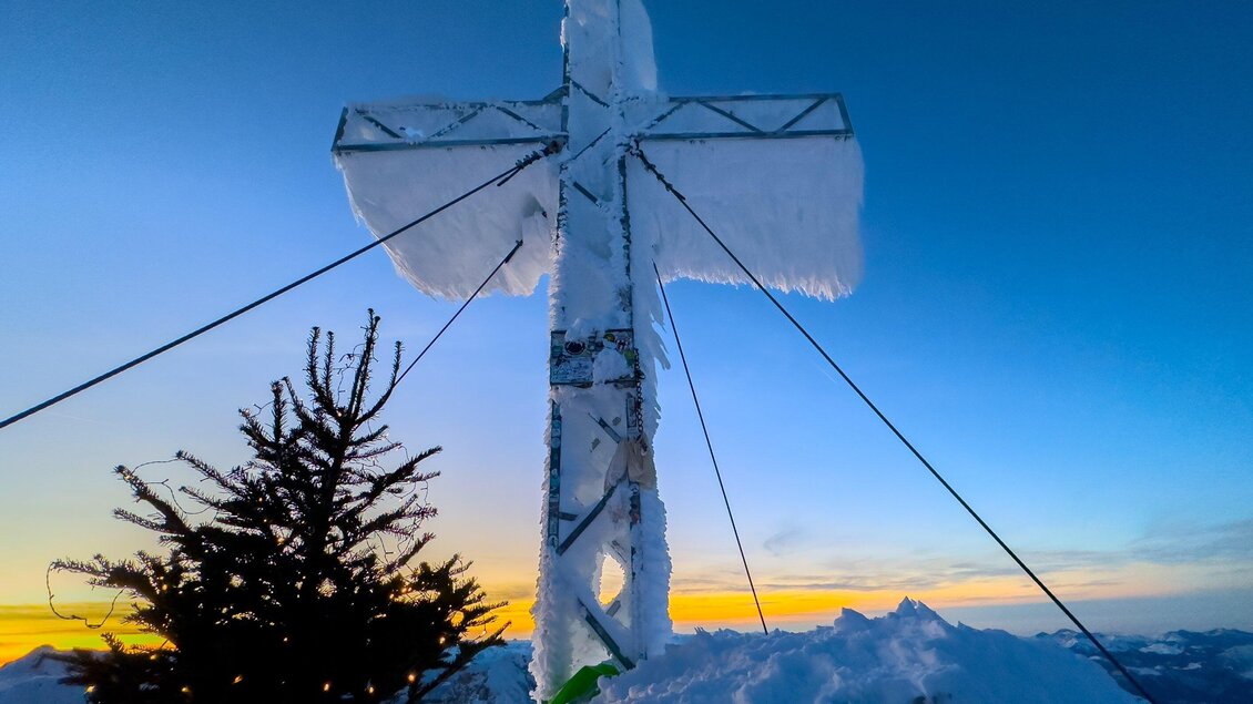 Ein großes Kreuz aus Metall, überzogen mit Schnee und Eis, steht auf einem schneebedeckten Gipfel. Im Hintergrund ist ein schöner Sonnenuntergang zu sehen. | © Paul Sodamin