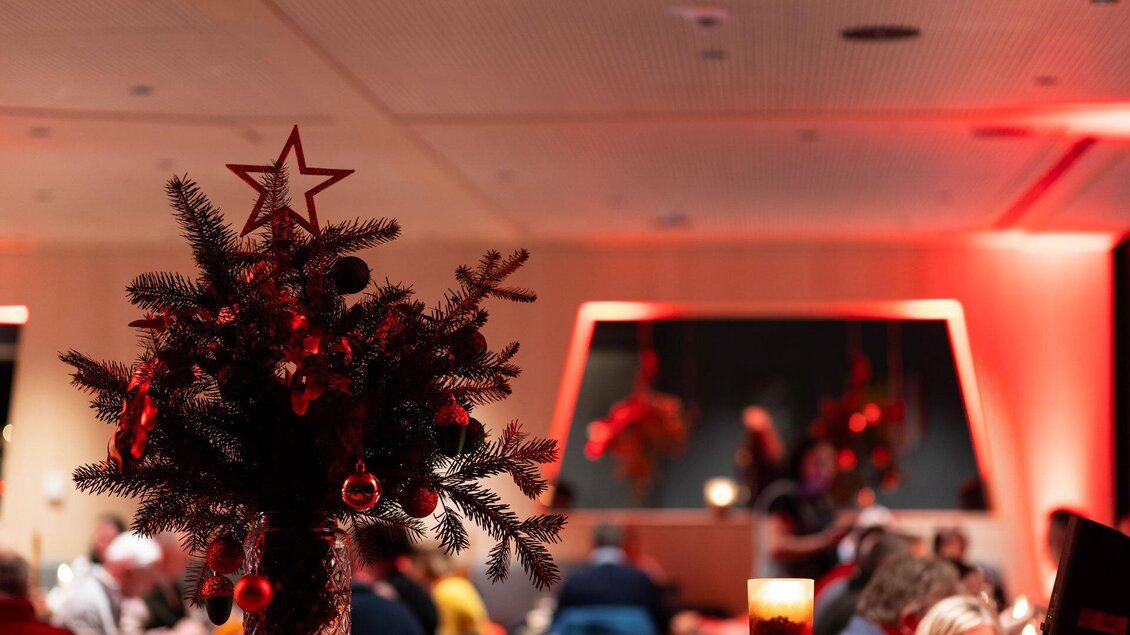 Ein dekorierter Weihnachtsbaum mit Stern und Schmuck steht im Vordergrund. Im Hintergrund sind unscharfe Menschen bei einer festlichen Veranstaltung zu sehen. | © Martin Huber