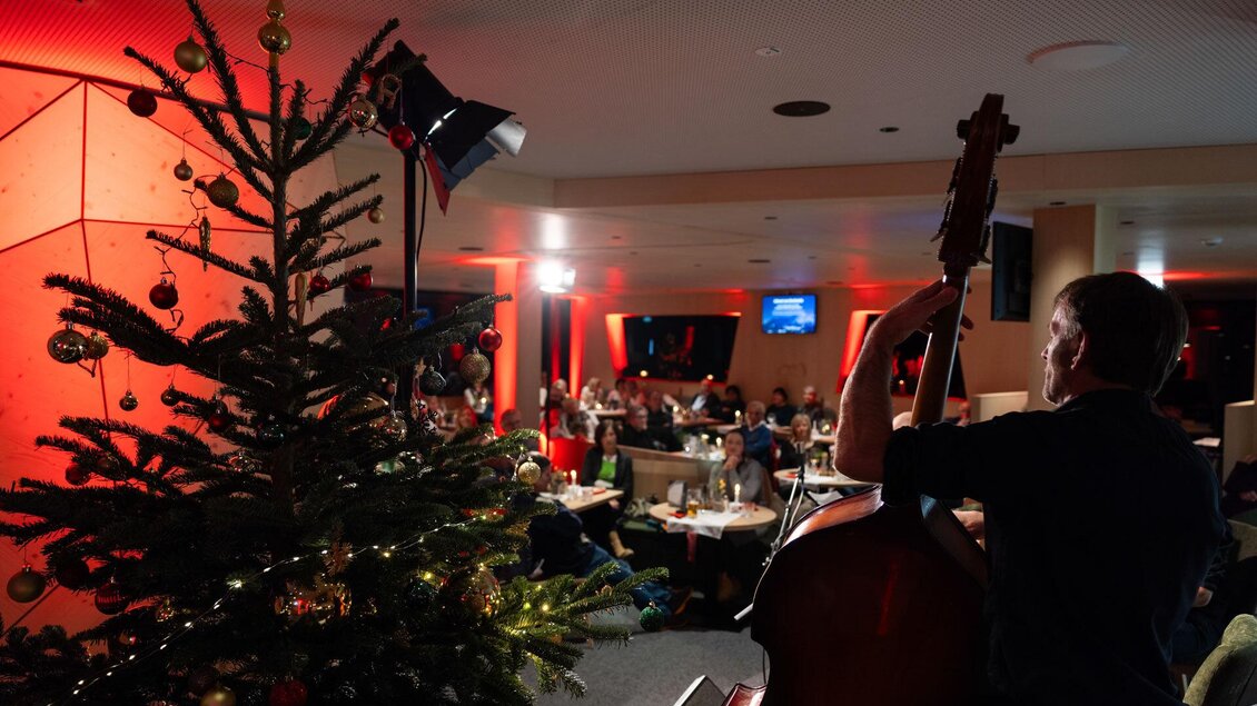 Eine festliche Weihnachtsfeier mit einem Musikanten, der Kontrabass spielt. Im Hintergrund steht ein dekorierter Weihnachtsbaum und Gäste sitzen an Tischen. | © Martin Huber