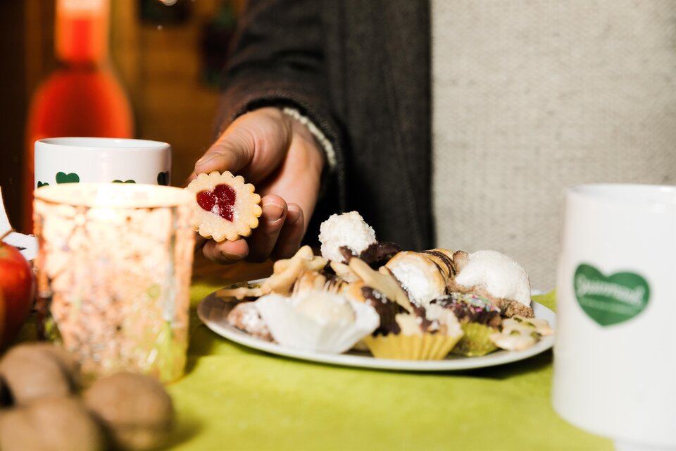 Ein Teller mit verschiedenen Süßigkeiten und Keksen wird von einer Hand gehalten. Im Hintergrund stehen zwei Tassen und eine Kerze auf einem Tisch. | © Lupi Spuma