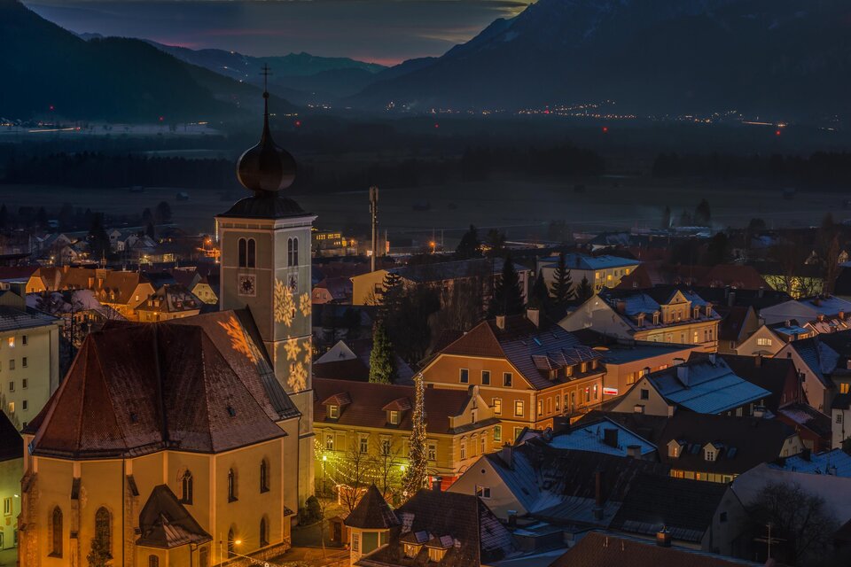 Eine malerische Stadtansicht bei Nacht mit beleuchteten Gebäuden und einer Kirche im Vordergrund. Im Hintergrund erheben sich majestätische Berge.