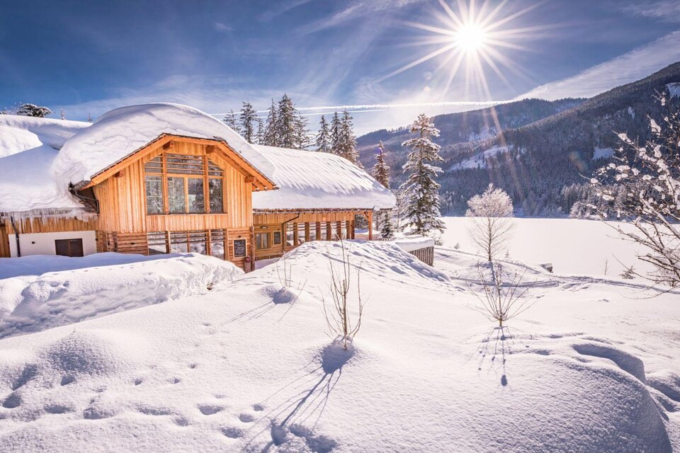 Ein wunderschönes Holzhaus in einer schneebedeckten Landschaft. Die Sonne strahlt über die Berge und schafft eine malerische Winterkulisse. | © Kohlröserlhütte_Genuss am See