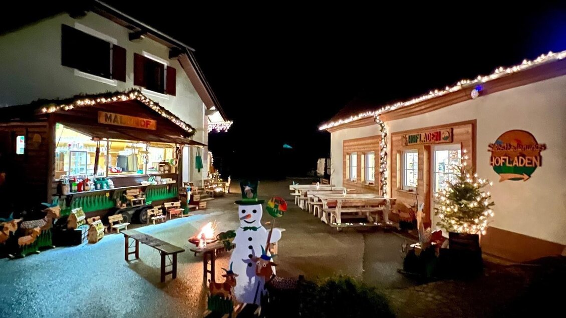 Ein festlich beleuchteter Hof mit zwei Gebäuden in der Nacht. Auf dem Weg stehen weihnachtliche Dekorationen und ein Schneemann. | © Malu