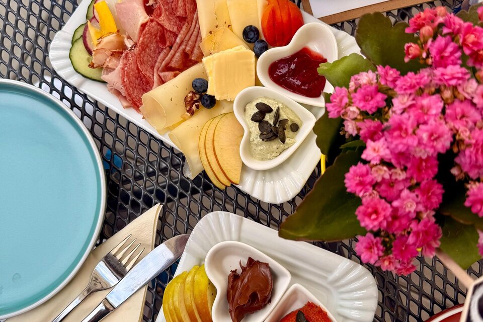 Ein appetitliches Frühstück mit Wurst, Käse, frischen Früchten und verschiedenen Dips. Die Teller sind auf einem dekorativen Tisch mit bunten Blumen angerichtet. | © Lisa-Marie Schiefer